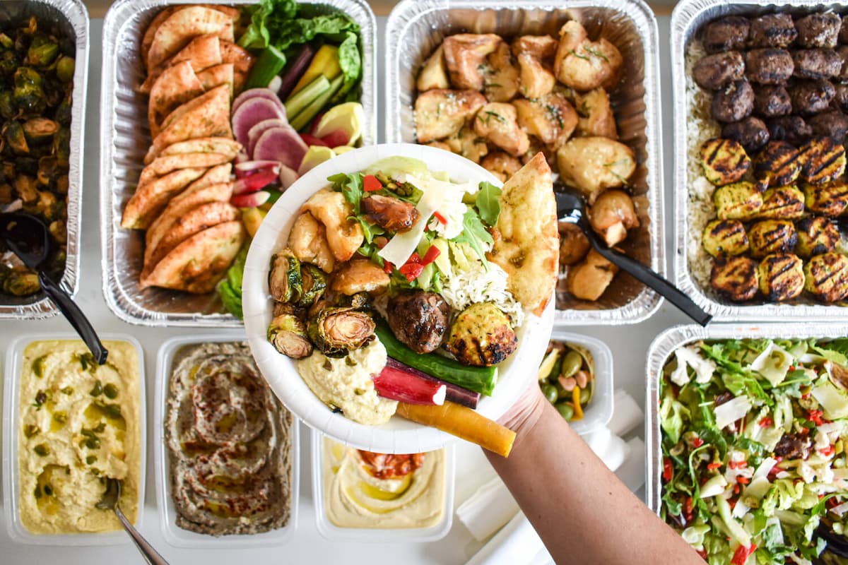 hand holding a plate of food available from Ema catering over trays of food packaged for catering